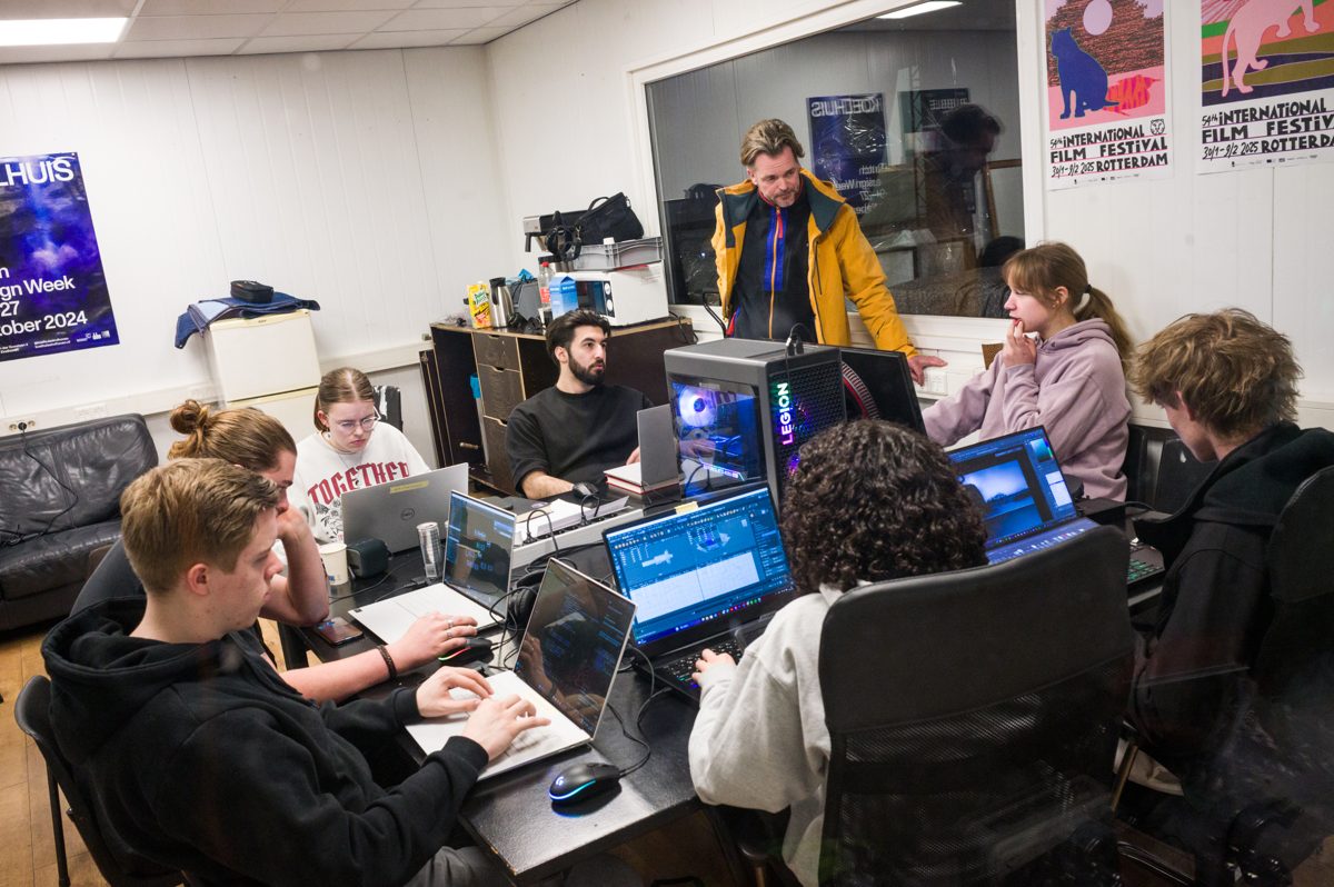 Studenten werken aan laptops rond een tafel terwijl een begeleider meekijkt, in een ruimte met IFFR-posters aan de muur.